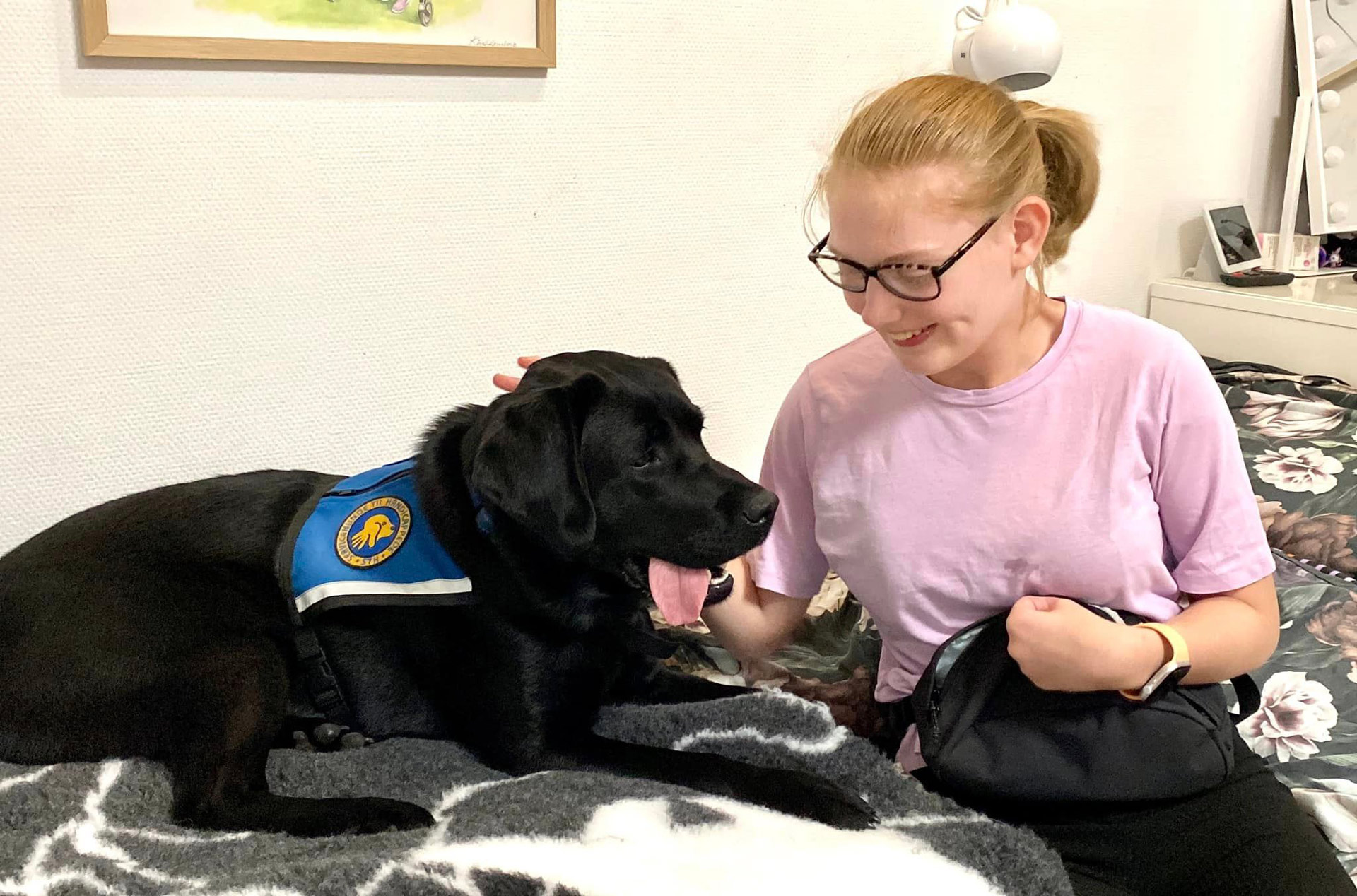 Marianne og servicehunden Stella der sidder i en seng sammen