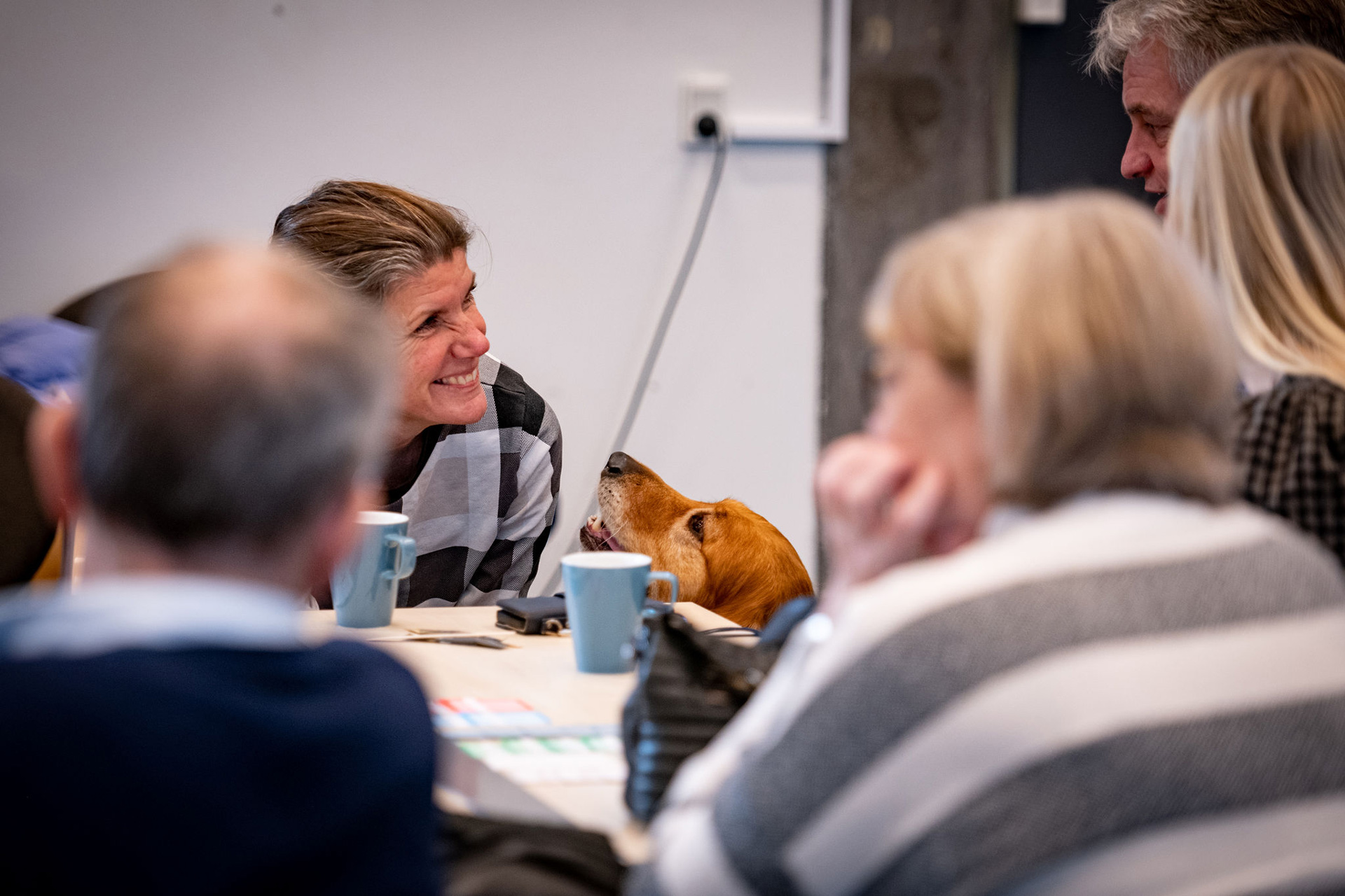En kvinder der griner og ved siden af står en servicehund der stikker hovedet op over et bord