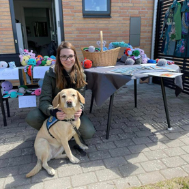 Emily sammen med hendes servicehund der står foran et bord med hæklede dyr til donation til STH