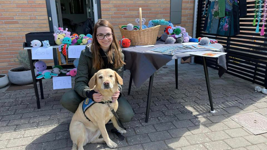 Emily sammen med hendes servicehund der står foran et bord med hæklede dyr til donation til STH